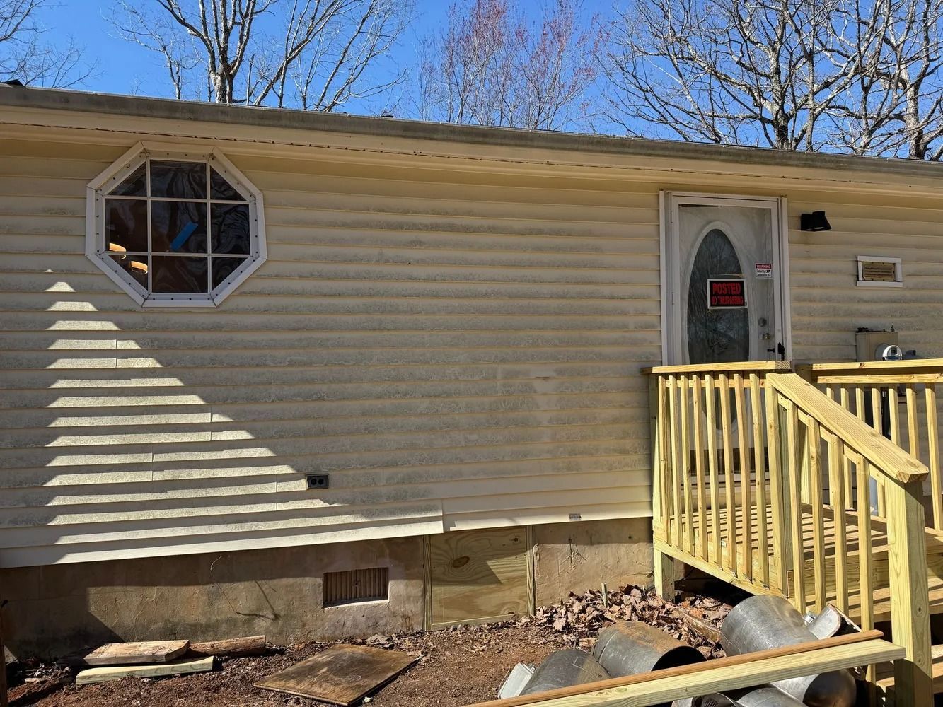 A beige mobile home with an octagonal window, a white door with a glass oval, and a wooden porch on a gravel lot.