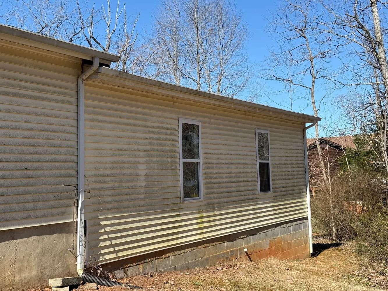 Side view of a light-colored, vinyl-sided home with two windows and a gutter downspout, set against a backdrop of trees.