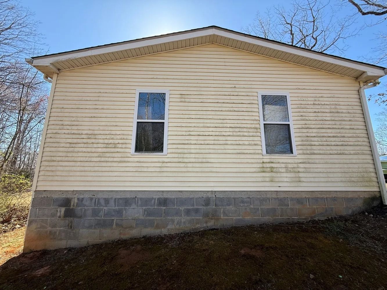Side view of a one-story house with yellow horizontal siding, two windows, and a concrete block foundation on a slope.