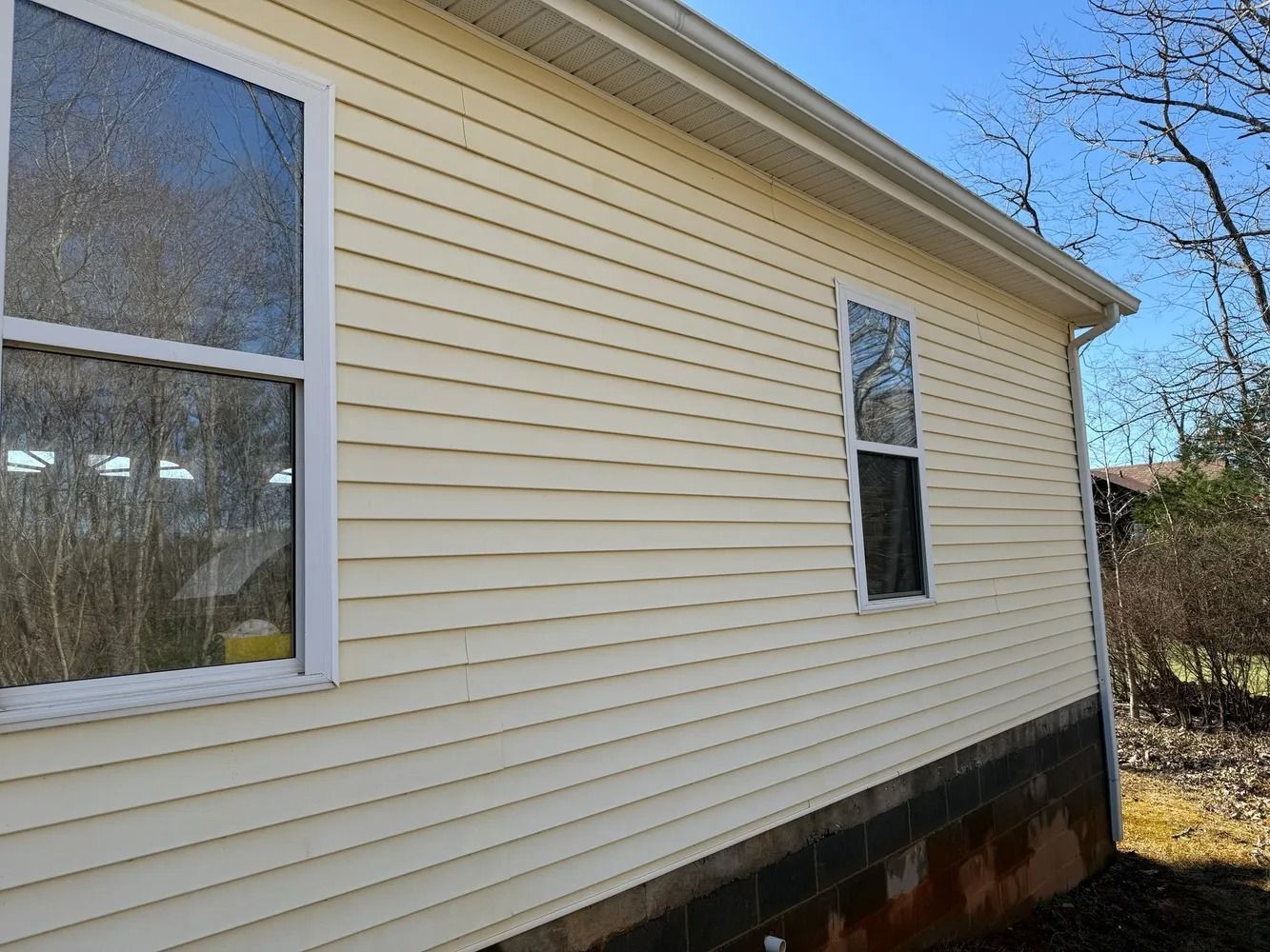 Exterior side of a house with pale yellow vinyl siding, two white-framed windows, and a dark stone foundation.