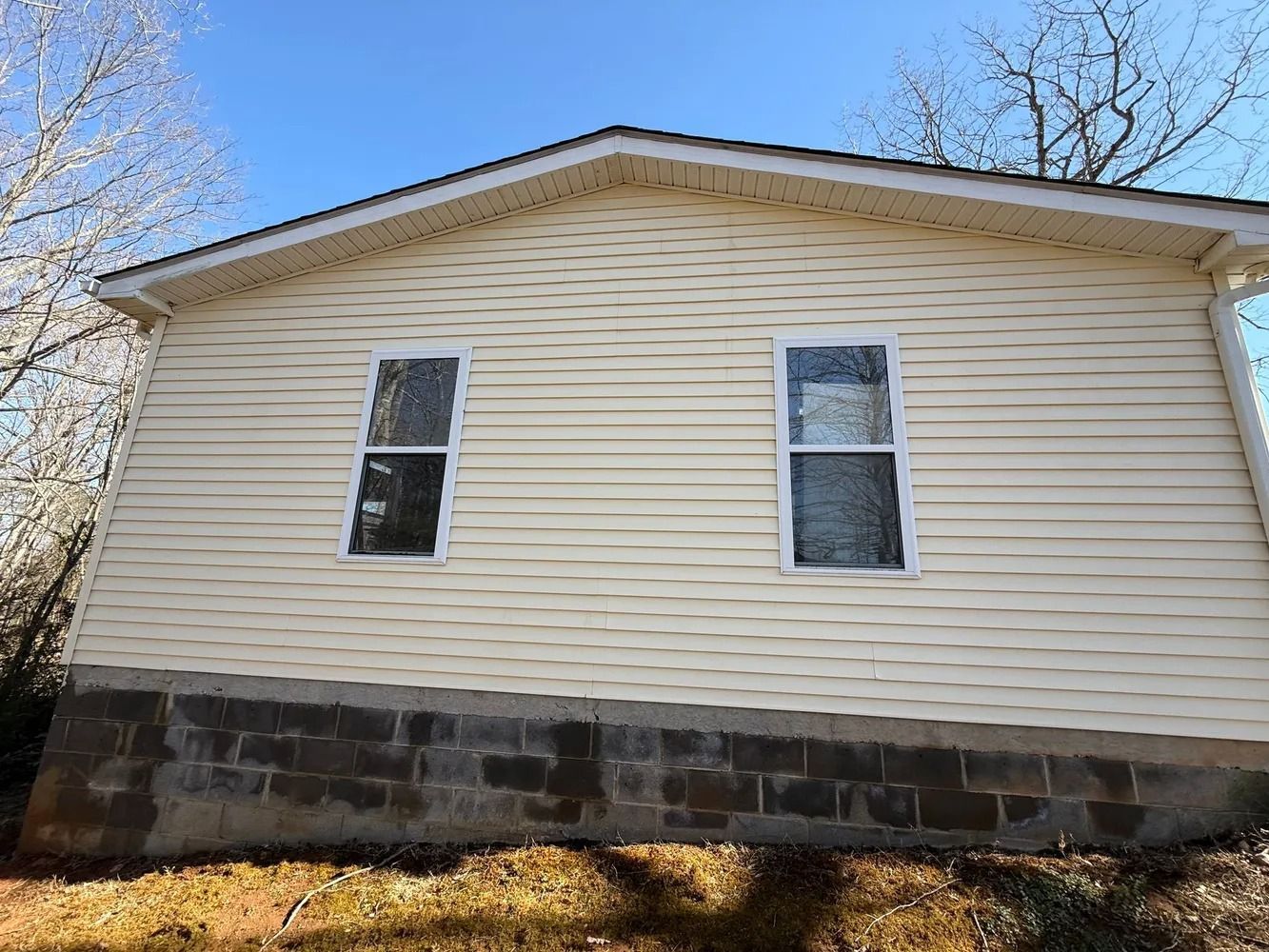 The side of a beige, vinyl-sided house with two windows, sitting on a dark concrete block foundation over a grassy slope.