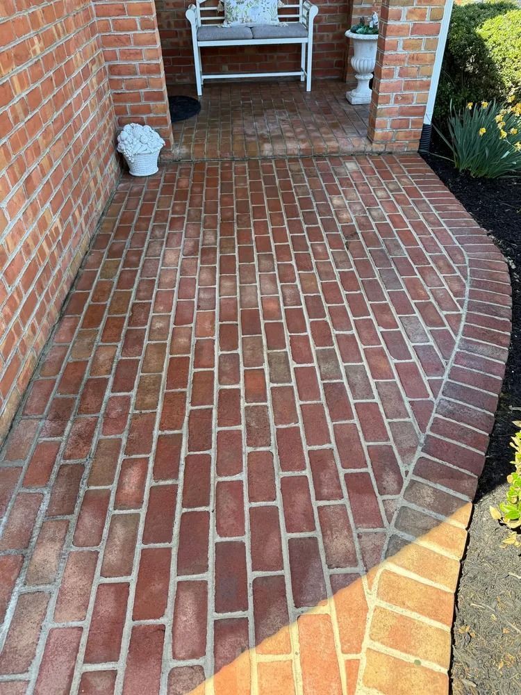 A curved brick walkway leading to a small front porch with a white bench and potted plants against a red brick house.