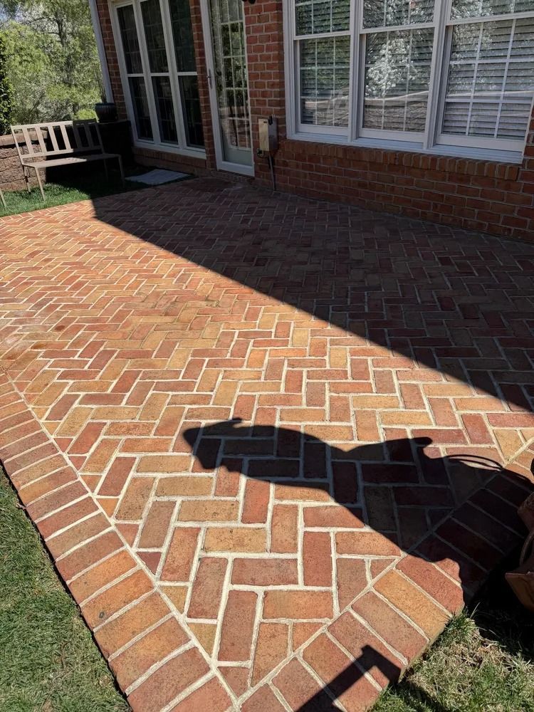 A brick patio with a herringbone pattern, bordered by a single row of bricks, beside a brick house and a metal bench.