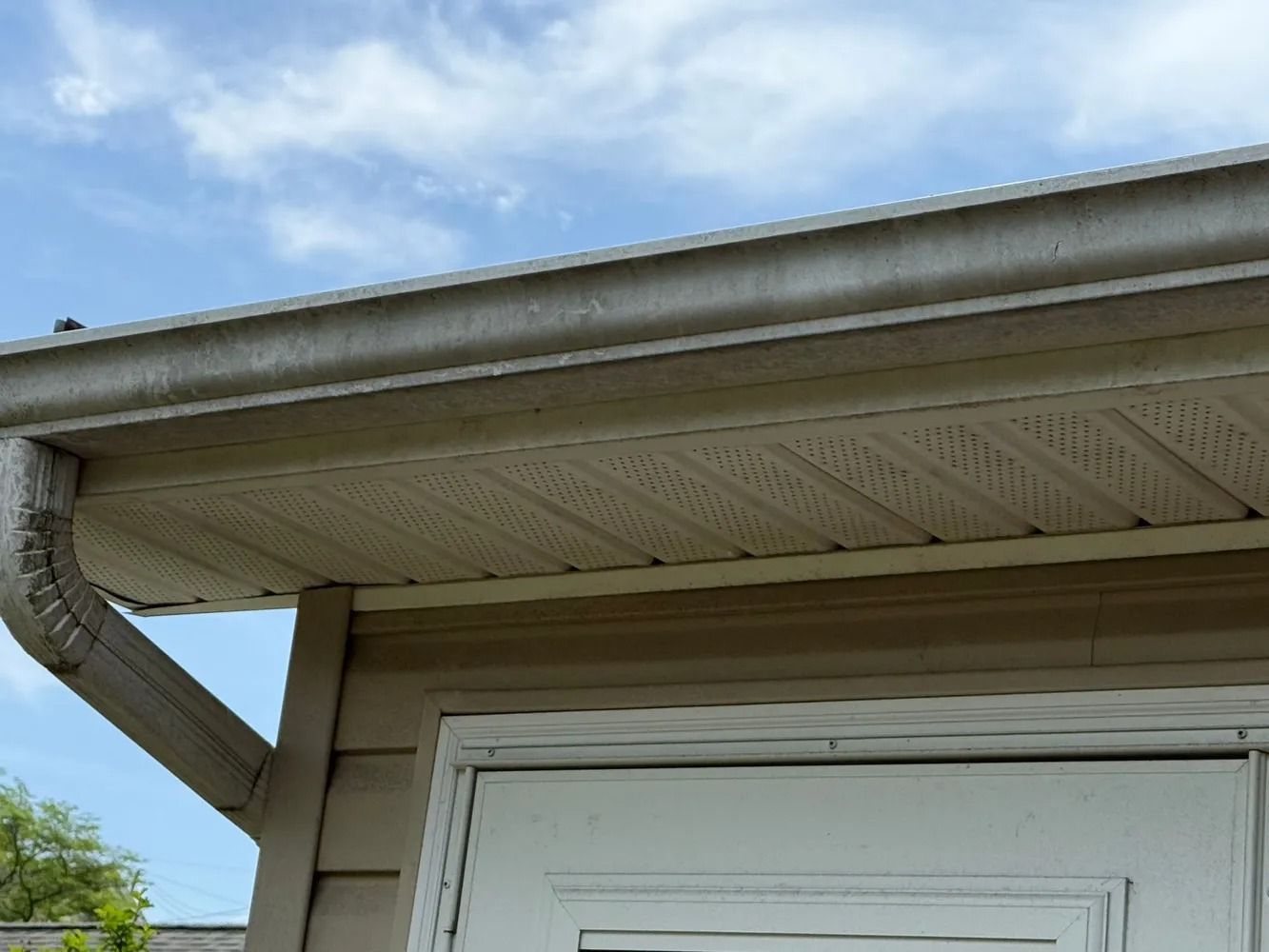 A low-angle view of a beige house exterior showing a tan gutter, perforated off-white soffit, and a white door frame.