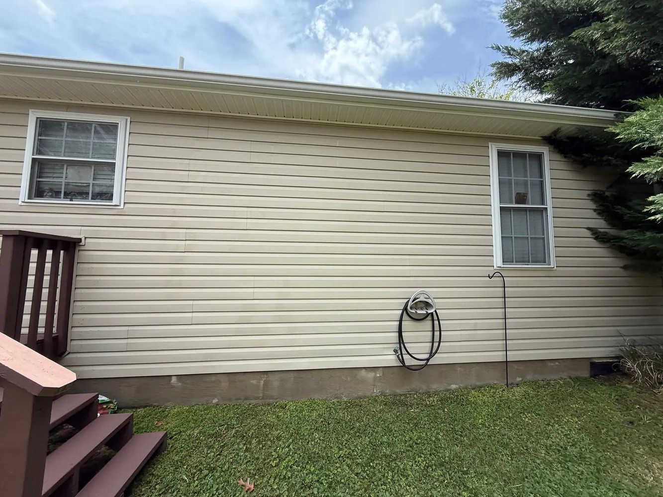 Tan vinyl siding on the exterior wall of a house with two windows, a wooden staircase, and a wall-mounted hose.