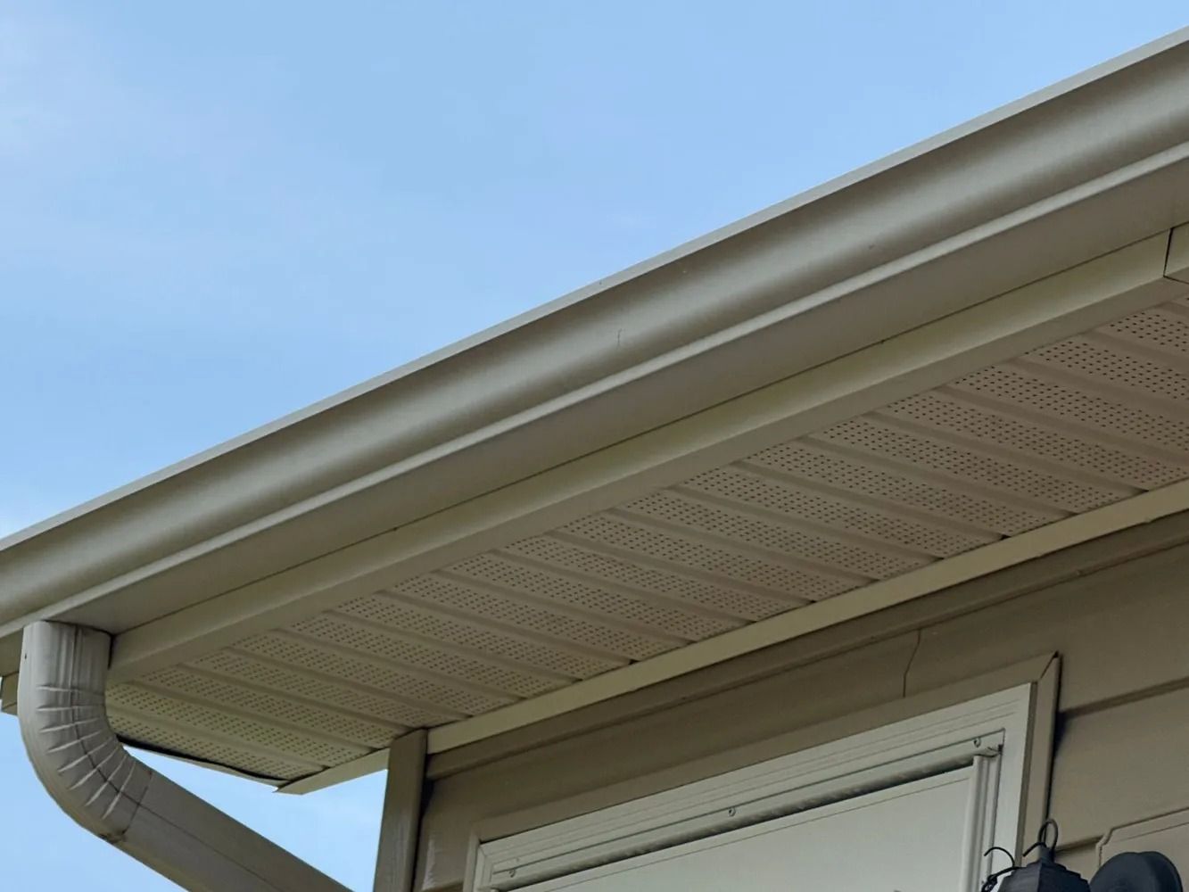 Beige house eaves, soffit, and gutter with an attached downspout against a clear blue sky.