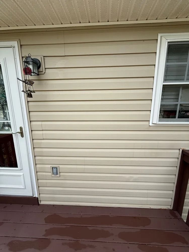 A tan horizontal vinyl-sided house exterior with a white door, a window, and a dark wooden deck.