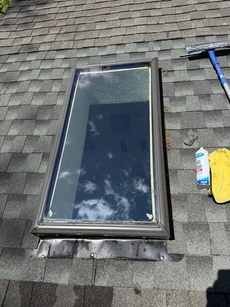 A rectangular skylight set into a gray asphalt shingle roof, with a cleaning squeegee, a spray bottle, and a cloth nearby.