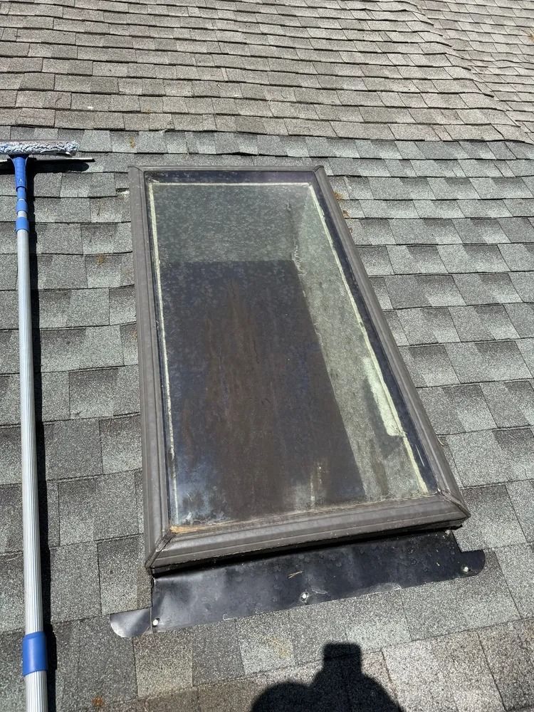 A rectangular skylight installed on a grey shingled roof, with an extension pole resting on the left side.