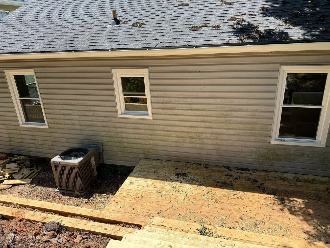 A side view of a house with tan siding, three white-framed windows, and an outdoor AC unit next to a wooden deck foundation.