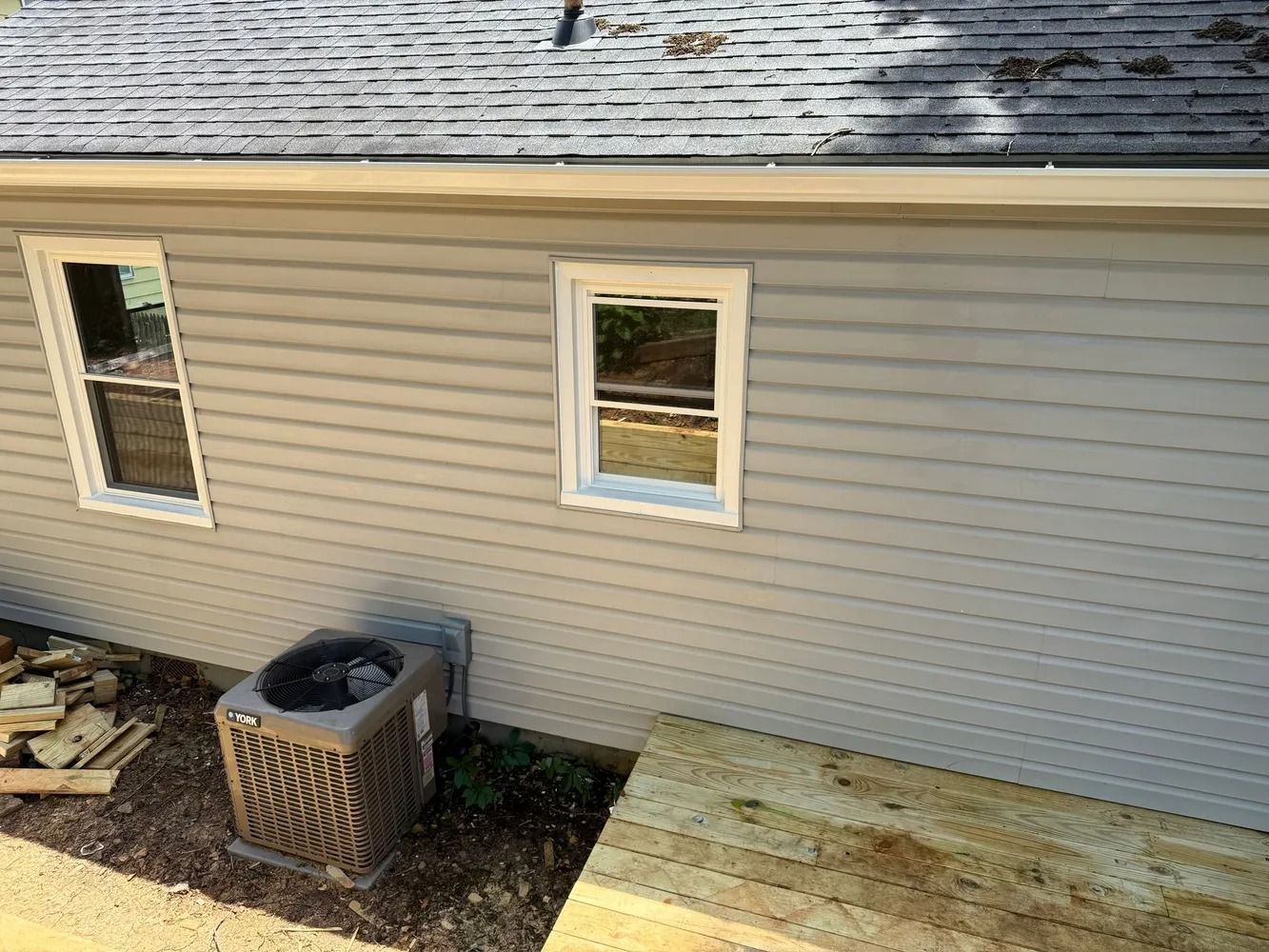 Light gray siding on a house wall featuring two windows, an exterior AC unit, and a newly built wooden deck in the front.
