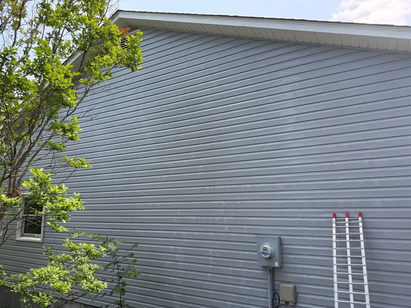 A light blue, horizontal vinyl-sided house wall with a small window, a utility meter, and a leaning metal ladder.