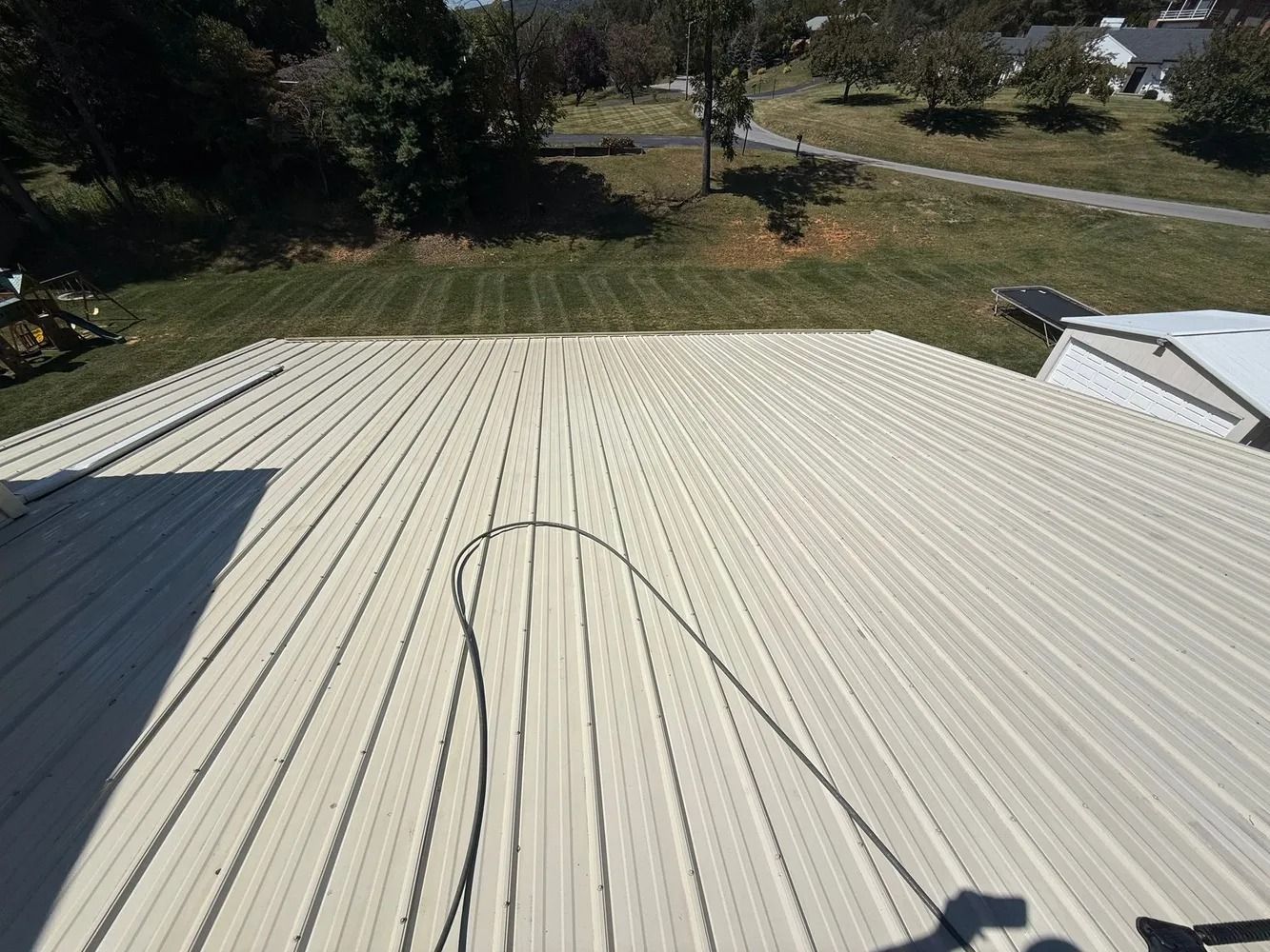 High-angle view of a beige metal roof sloping downward toward a grassy yard under a clear blue sky.