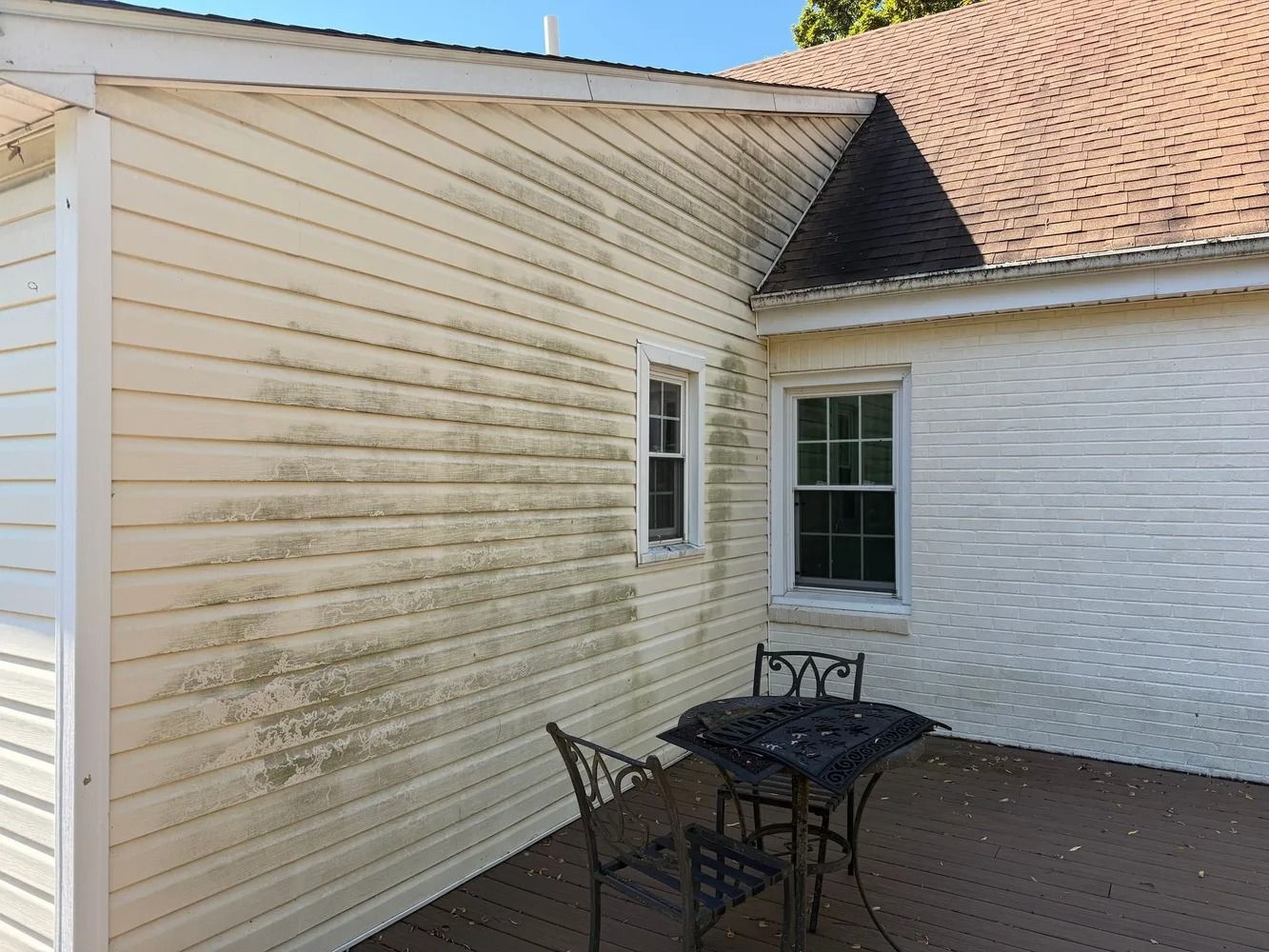 Cream-colored vinyl siding on a house wall showing significant green algae growth next to a deck with outdoor furniture.