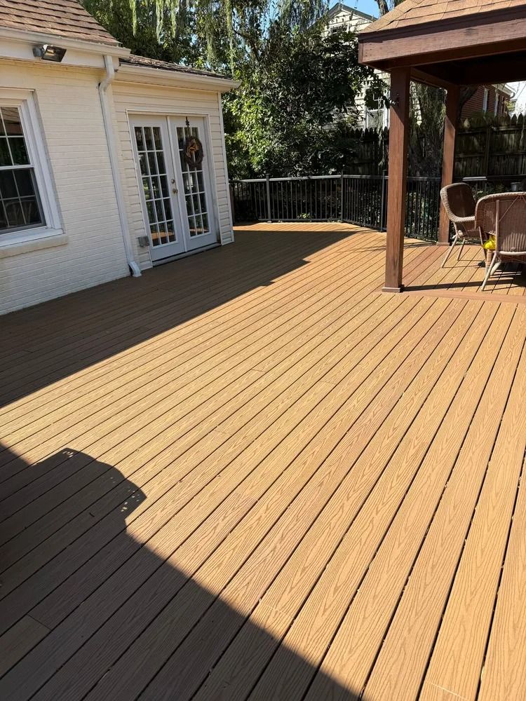 A wooden deck with a beige surface, leading to a house with white double doors and a partial outdoor roof structure.