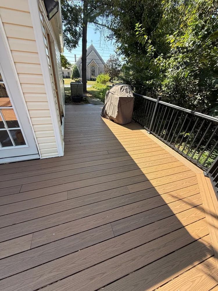A sunny outdoor deck with brown wooden planks, a black metal railing, and a covered grill near a house exterior.