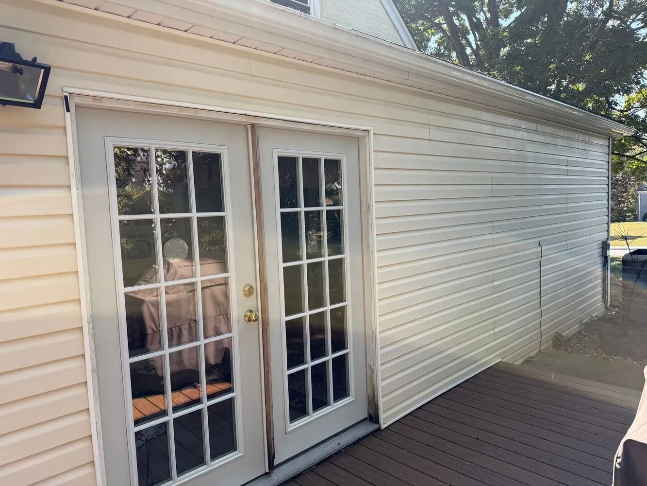Exterior view of a house wall with beige vinyl siding and a set of white French doors leading onto a wooden deck.