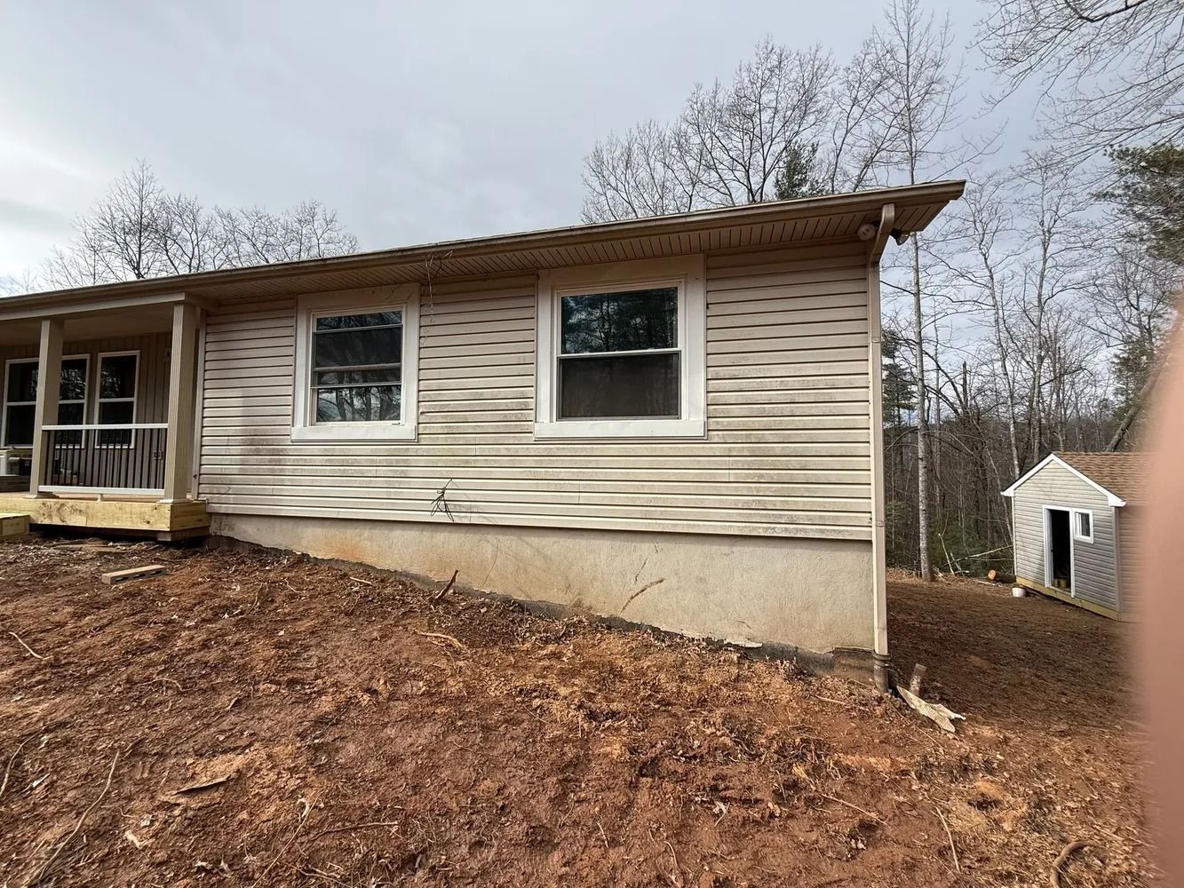 A beige, single-story house with wavy siding sits on a sloped dirt lot next to a small shed.