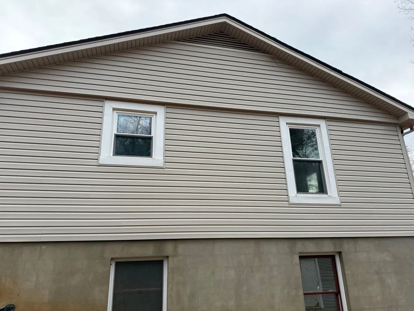A tan vinyl-sided house exterior with two upper-story windows and two basement windows against a cloudy sky.