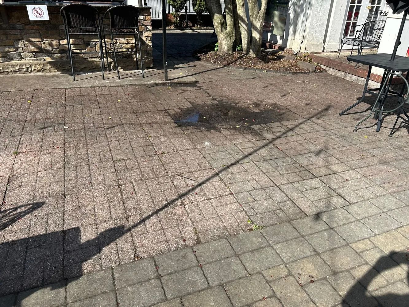 An outdoor patio area with dark brick pavers, a small puddle, patio furniture, and the long shadow of a person.