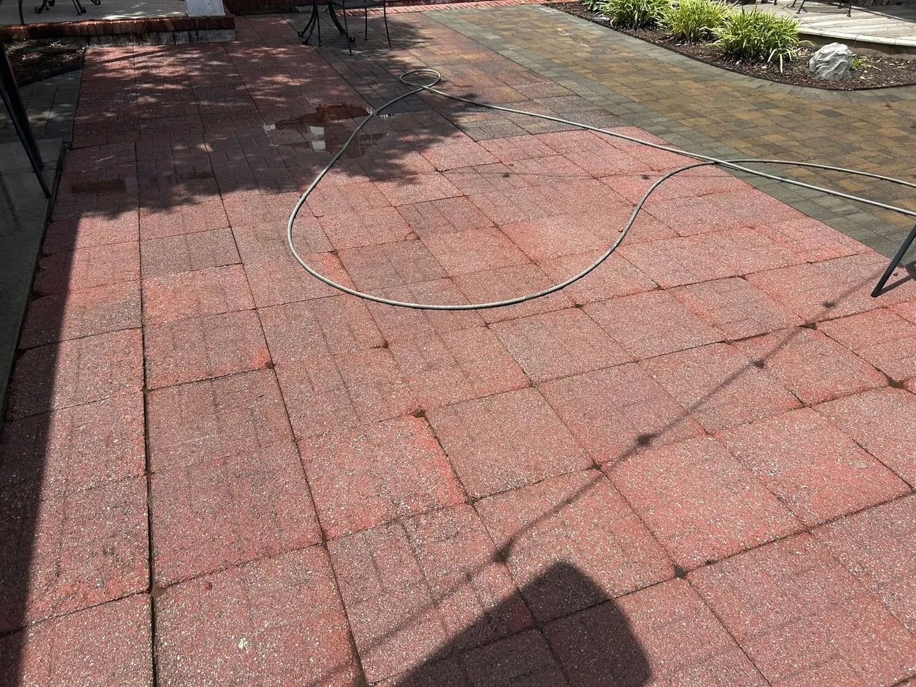 A patio covered in rectangular, reddish-brown pavers with a garden hose lying across the surface.