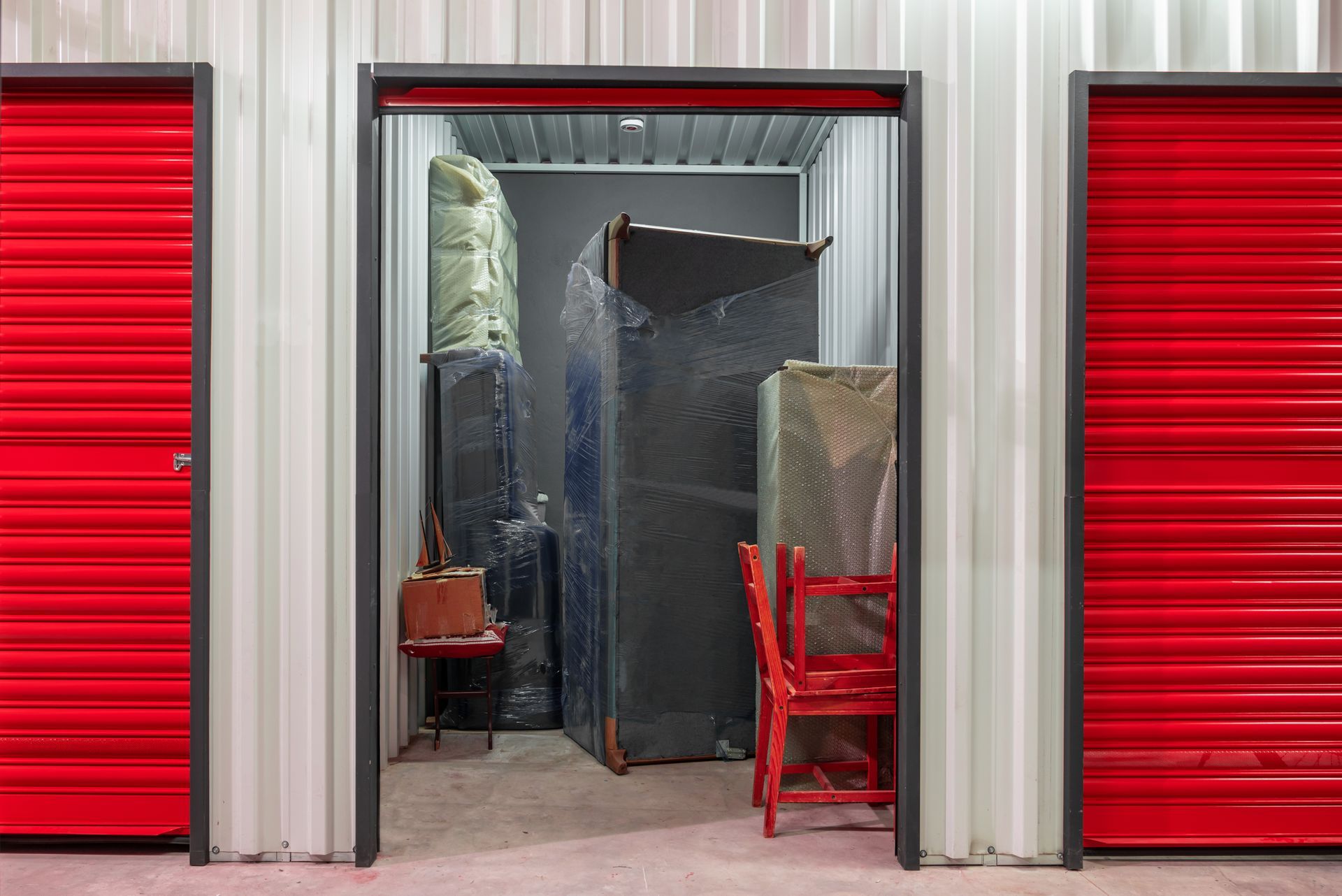 Storage unit with red door open, revealing furniture wrapped for protection, among other items.