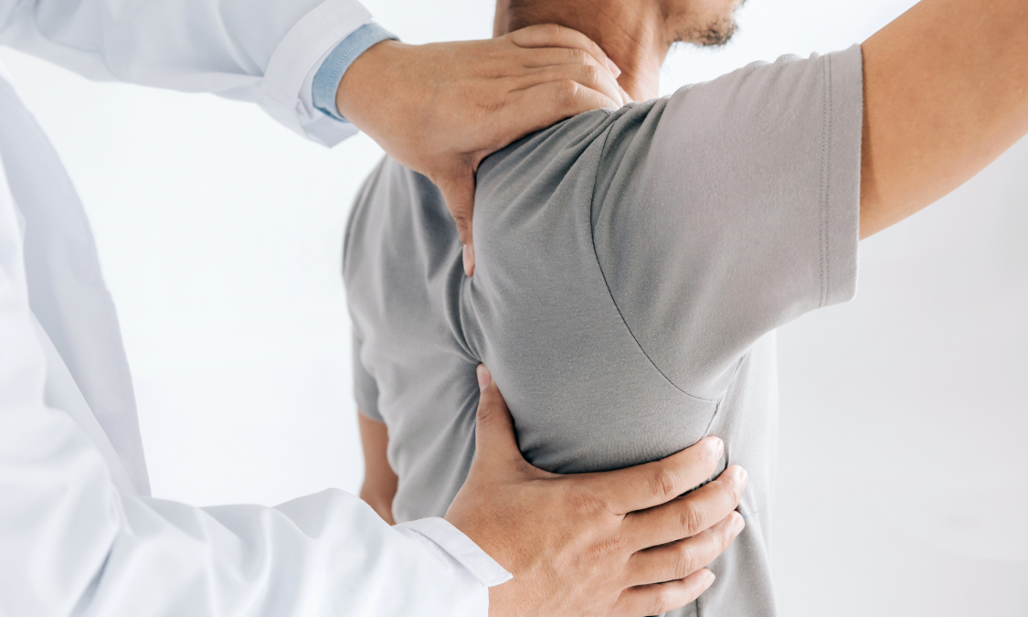Doctor examining patient's shoulder and back, both wearing light-colored shirts. White background.