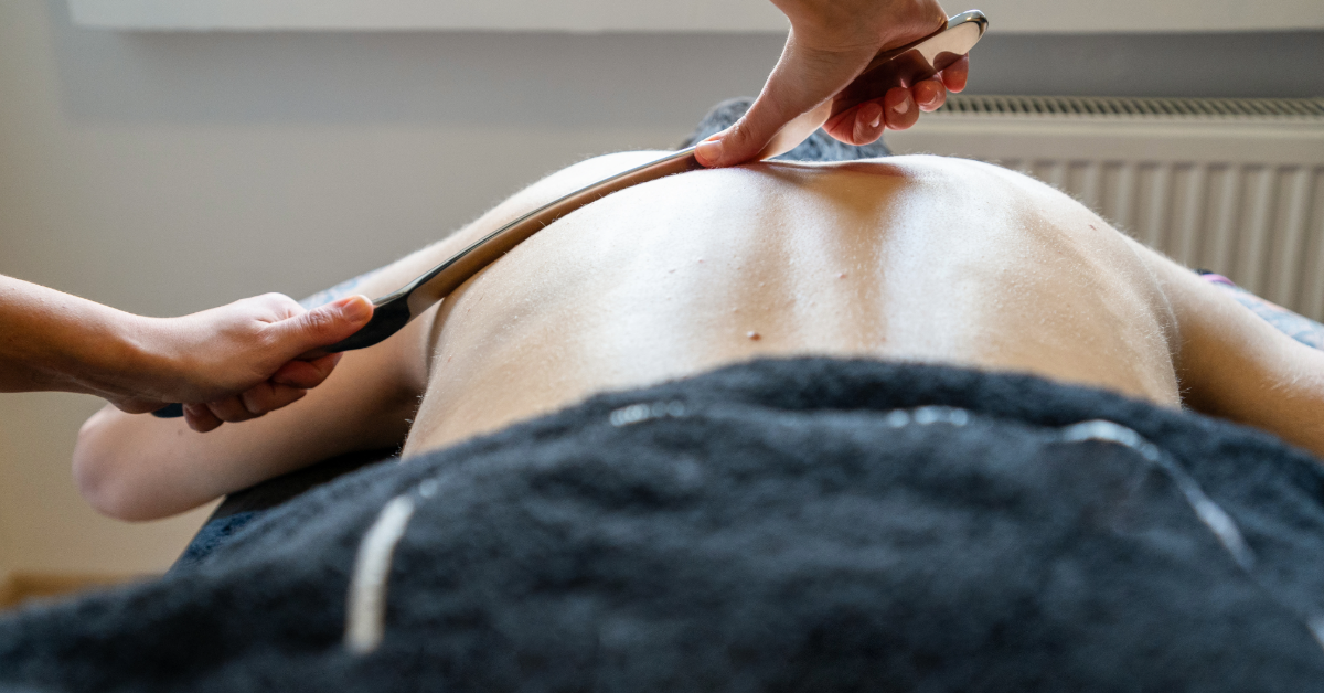 Person receiving Gua Sha massage on their back, held by two hands, a dark towel beneath.