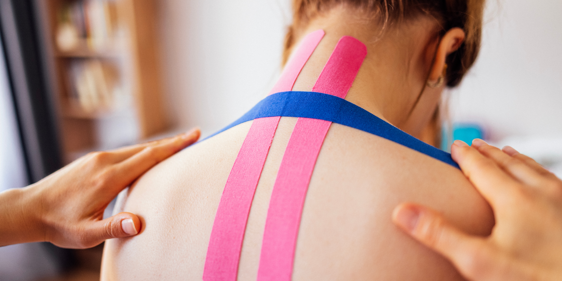 Person's back and neck with pink and blue kinesiology tape, being touched by hands.