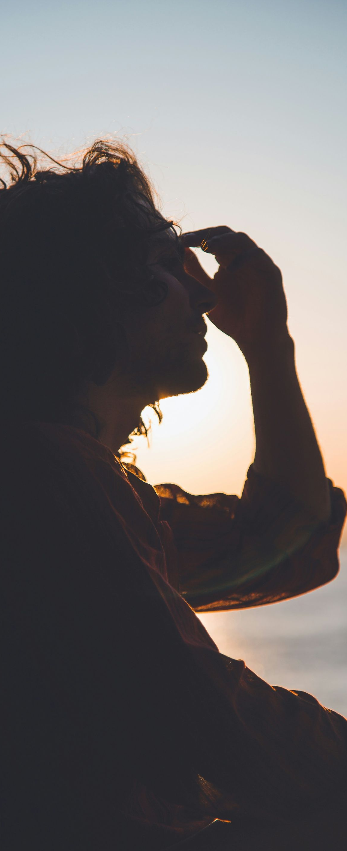 Silhouette of person with curly hair, hand on forehead, looking out at ocean during sunset.