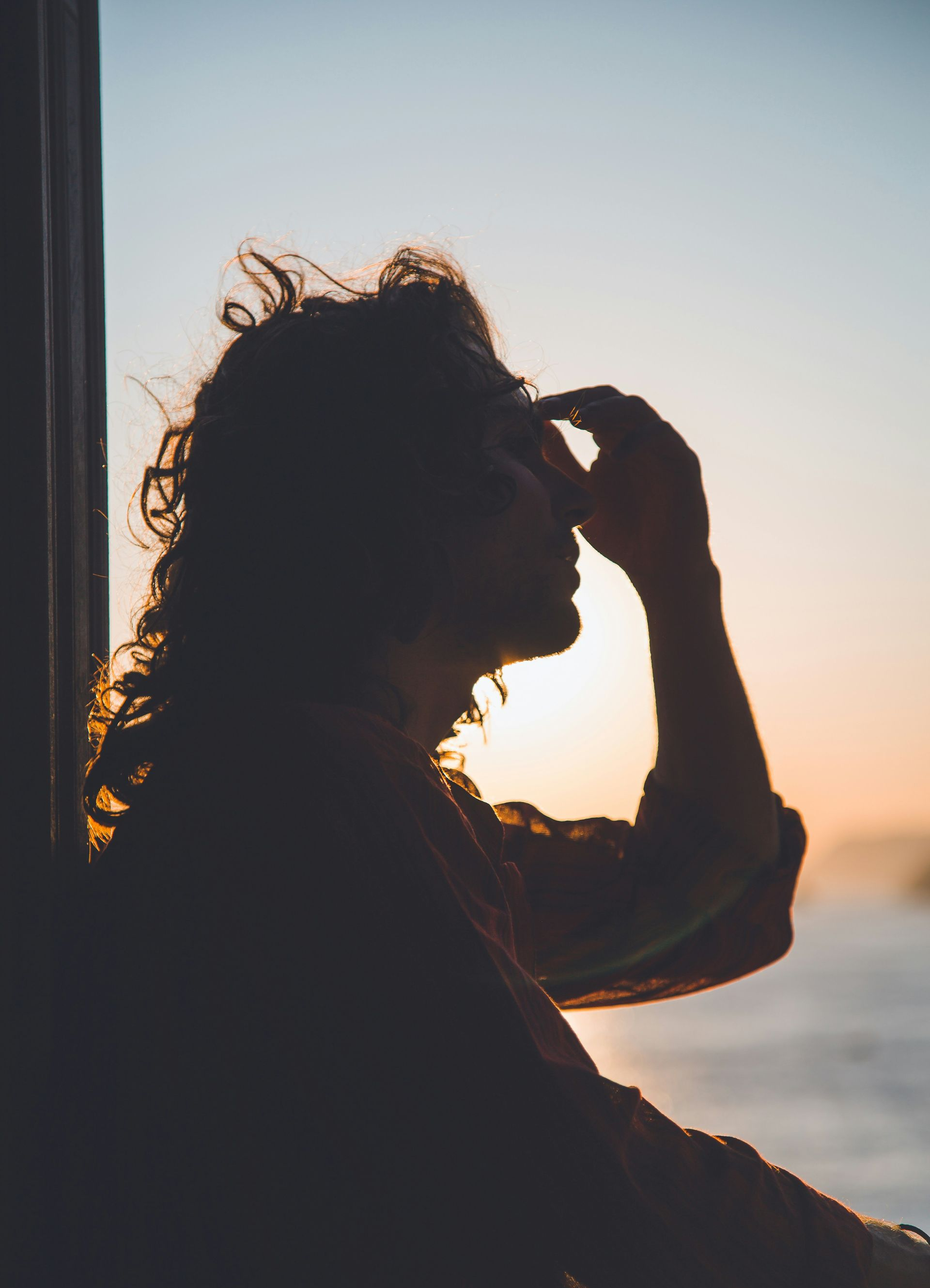 Silhouette of a person with curly hair, hand on forehead, looking out at a sunset over water.