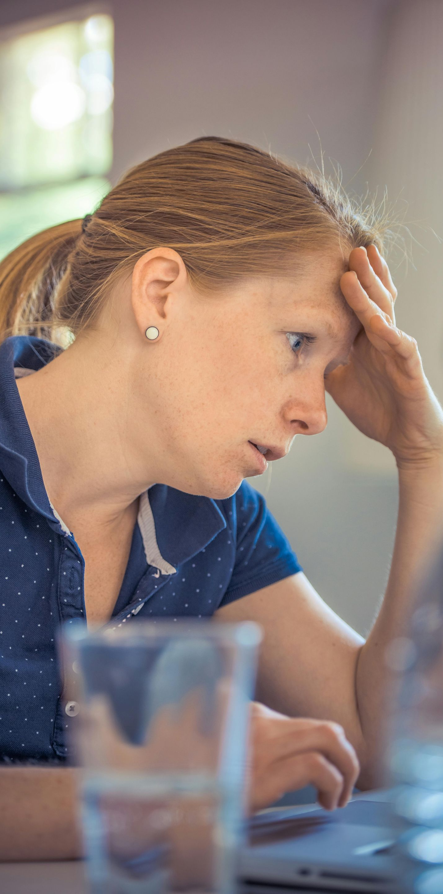 Woman with light hair, stressed expression, hand on forehead, looking at laptop.