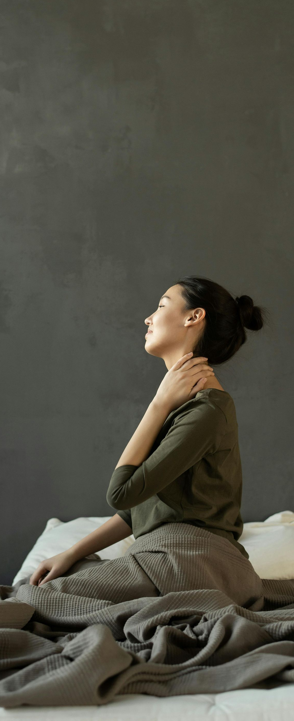 Woman sitting on a bed, hand on her shoulder. She looks up, eyes closed, with a gray wall in the background.