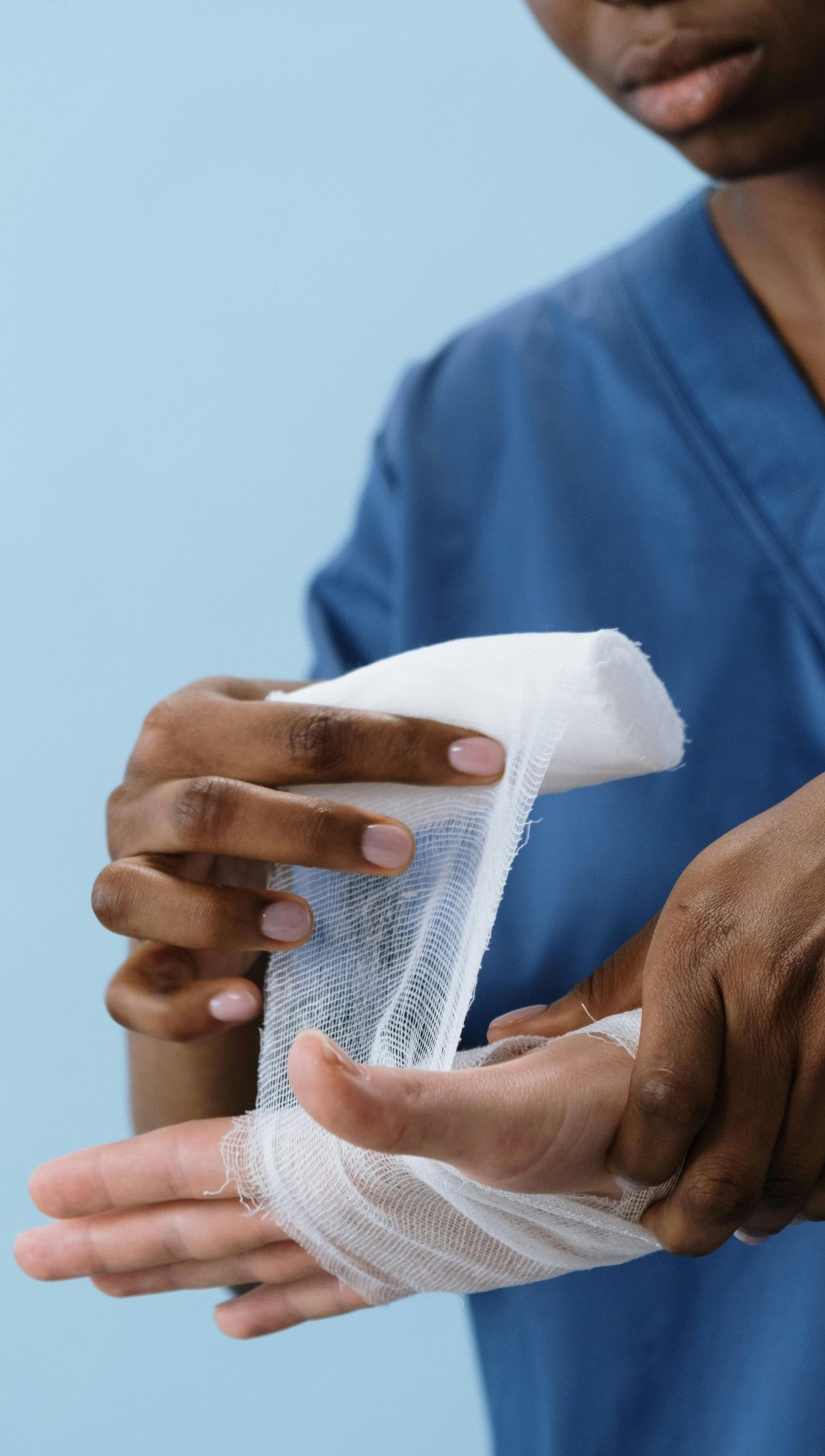 Person wrapping a white bandage around a hand, blue scrubs and background.