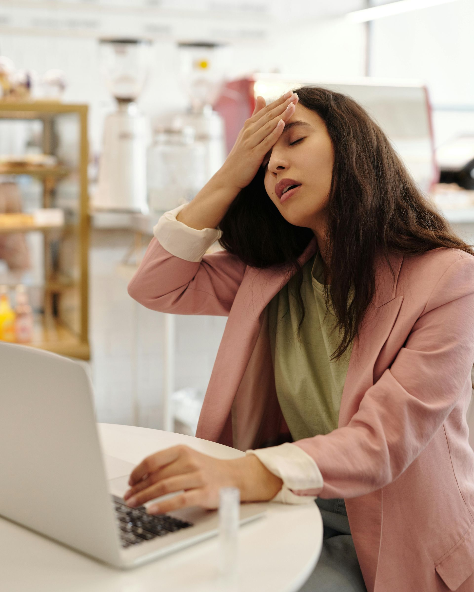 Woman with hand on forehead, looking stressed at a laptop in a cafe.