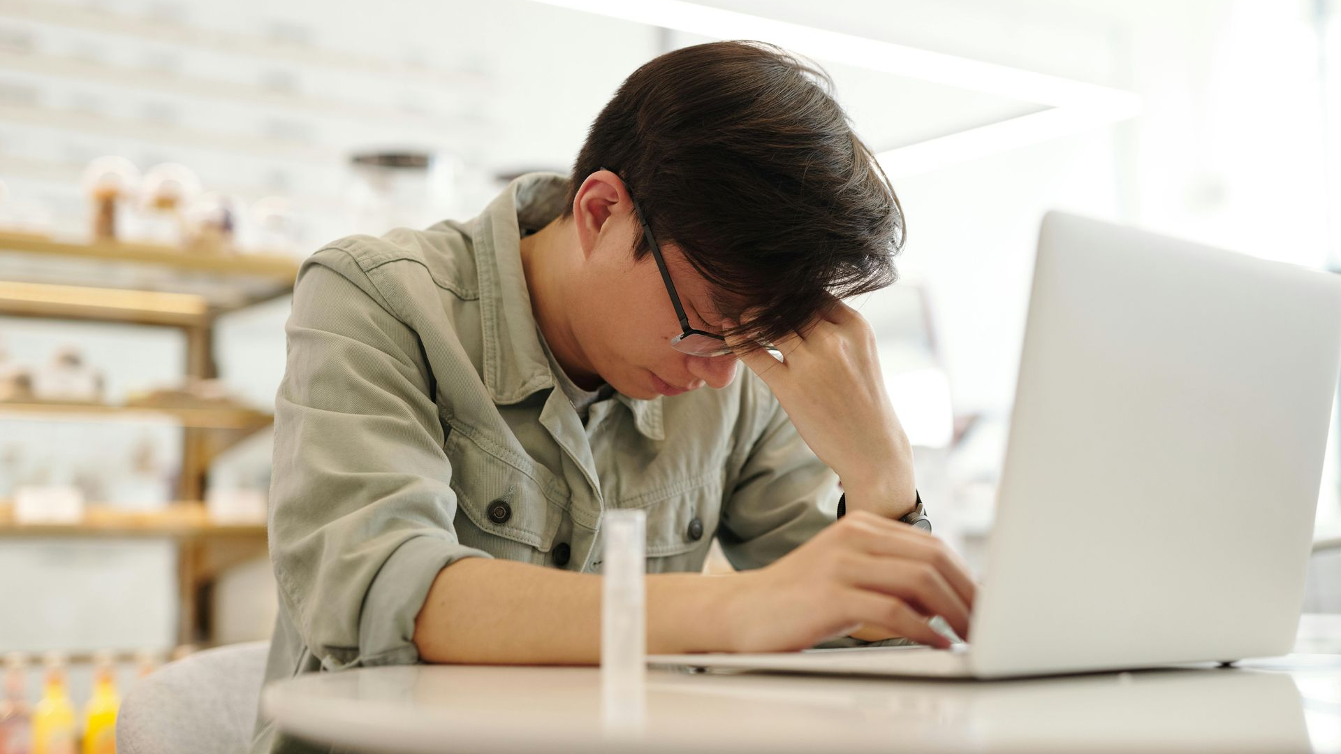 Person with glasses, head in hand, seated at a table with a laptop, looking stressed.