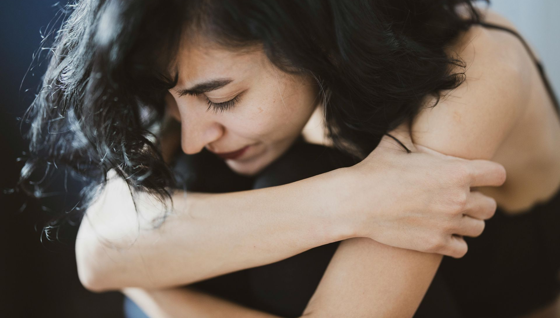Woman with dark hair, eyes closed, hugs knees, looking down.