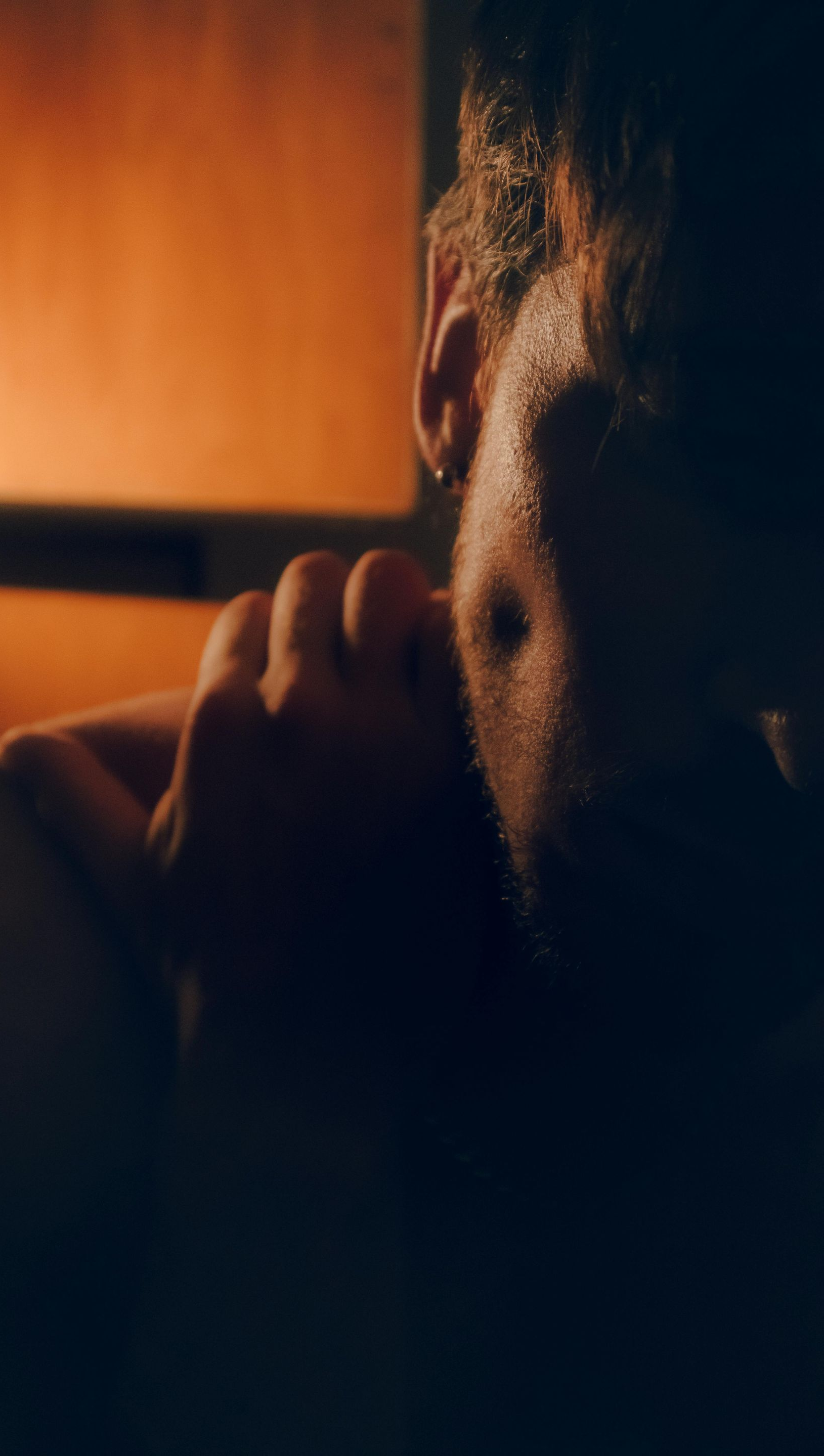 A person with curly hair, partially obscured, holding a hand near their face, lit by warm orange light.