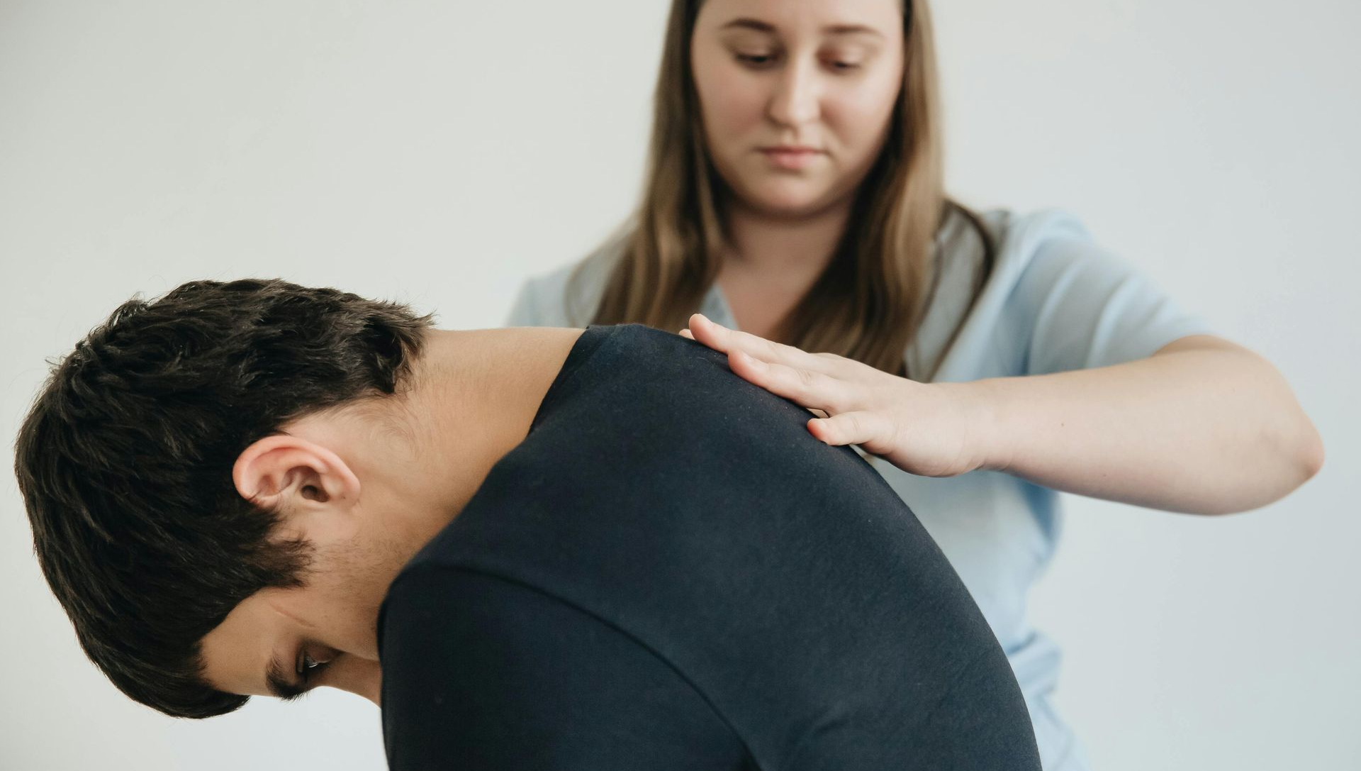 Person with dark hair bent forward as another person supports their back; inside setting, white background.