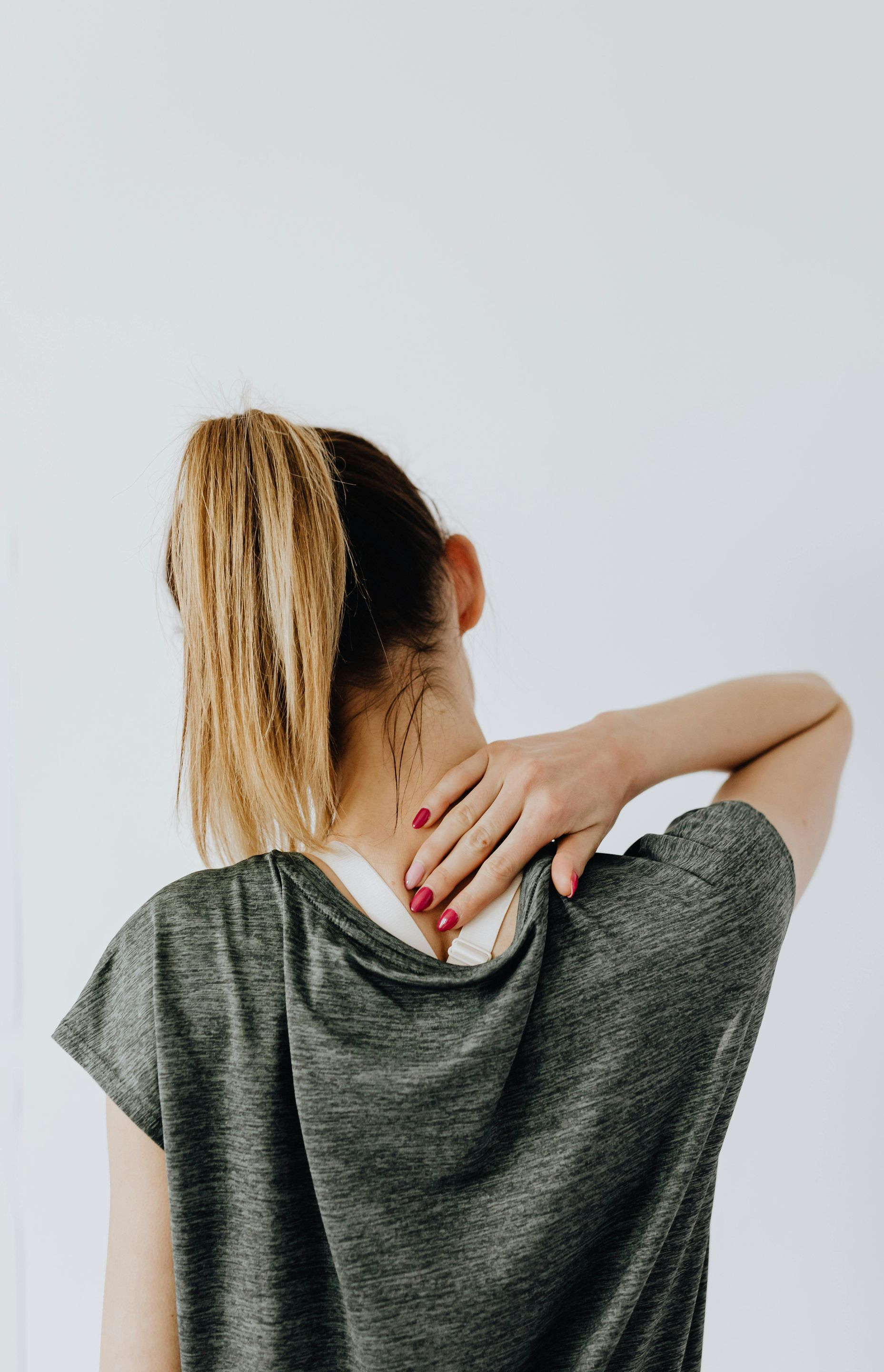 Woman touching her neck, possibly experiencing pain. Wearing gray shirt, light hair in a ponytail against a white background.