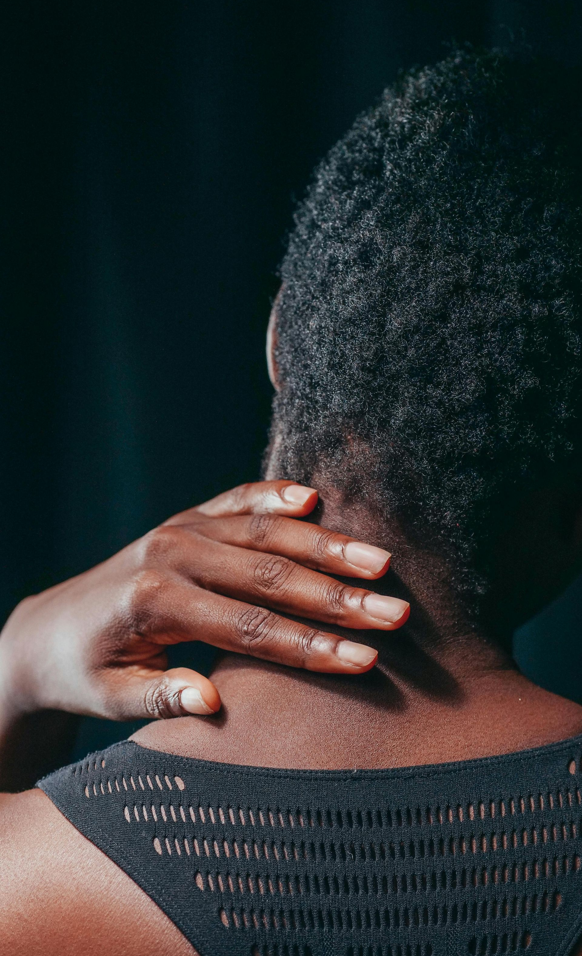 Person's hand on their neck, likely massaging it. Back view, dark skin, short, textured hair, against a dark backdrop.