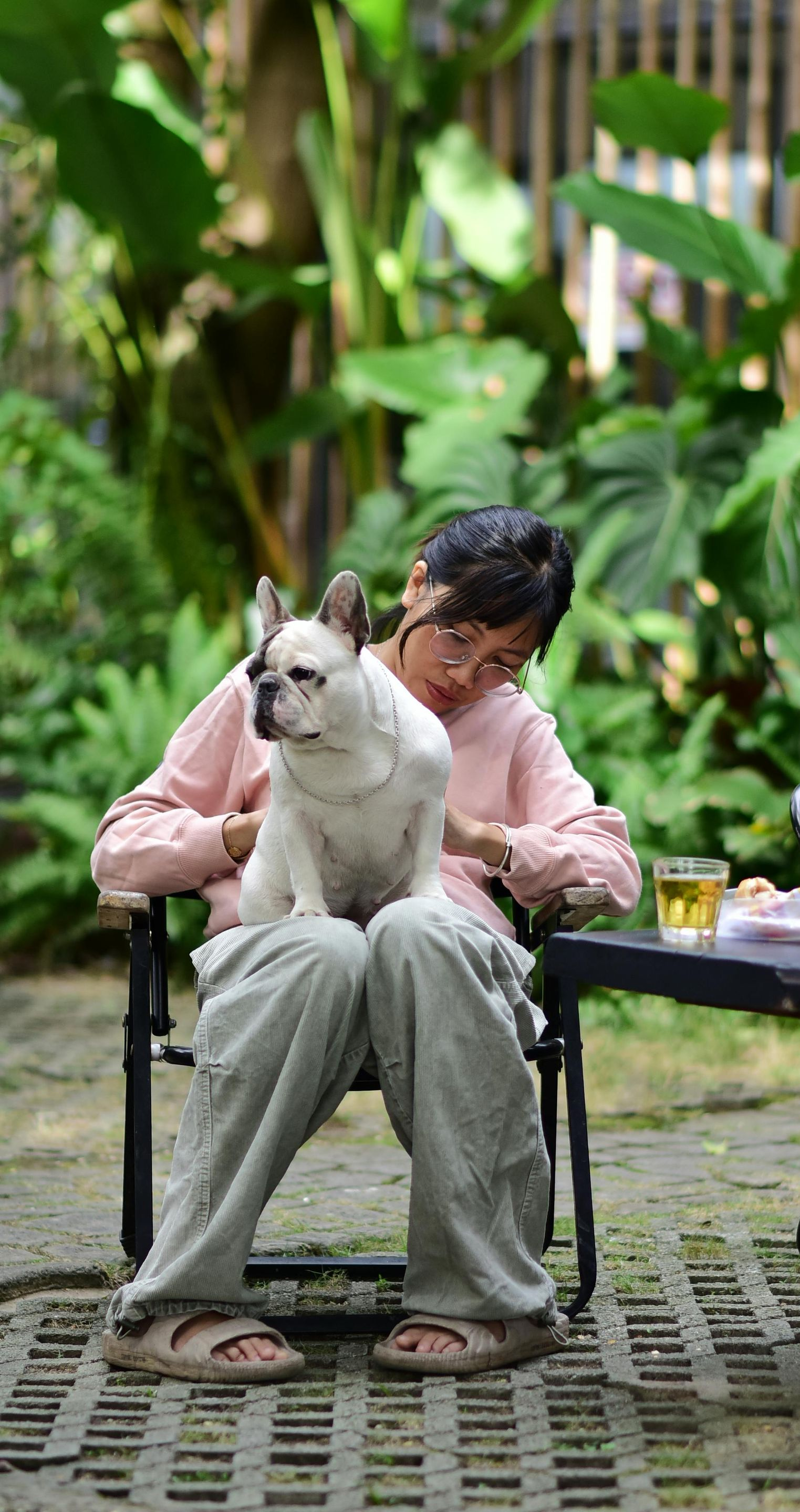 Person seated, dog on lap, outdoors. Dog is white and tan, person is wearing pink shirt and green pants.