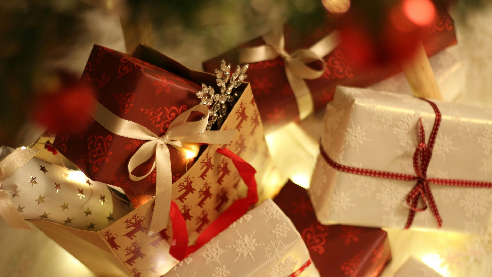 Pile of wrapped Christmas gifts in red, white, and patterned paper, tied with ribbons, with string lights.