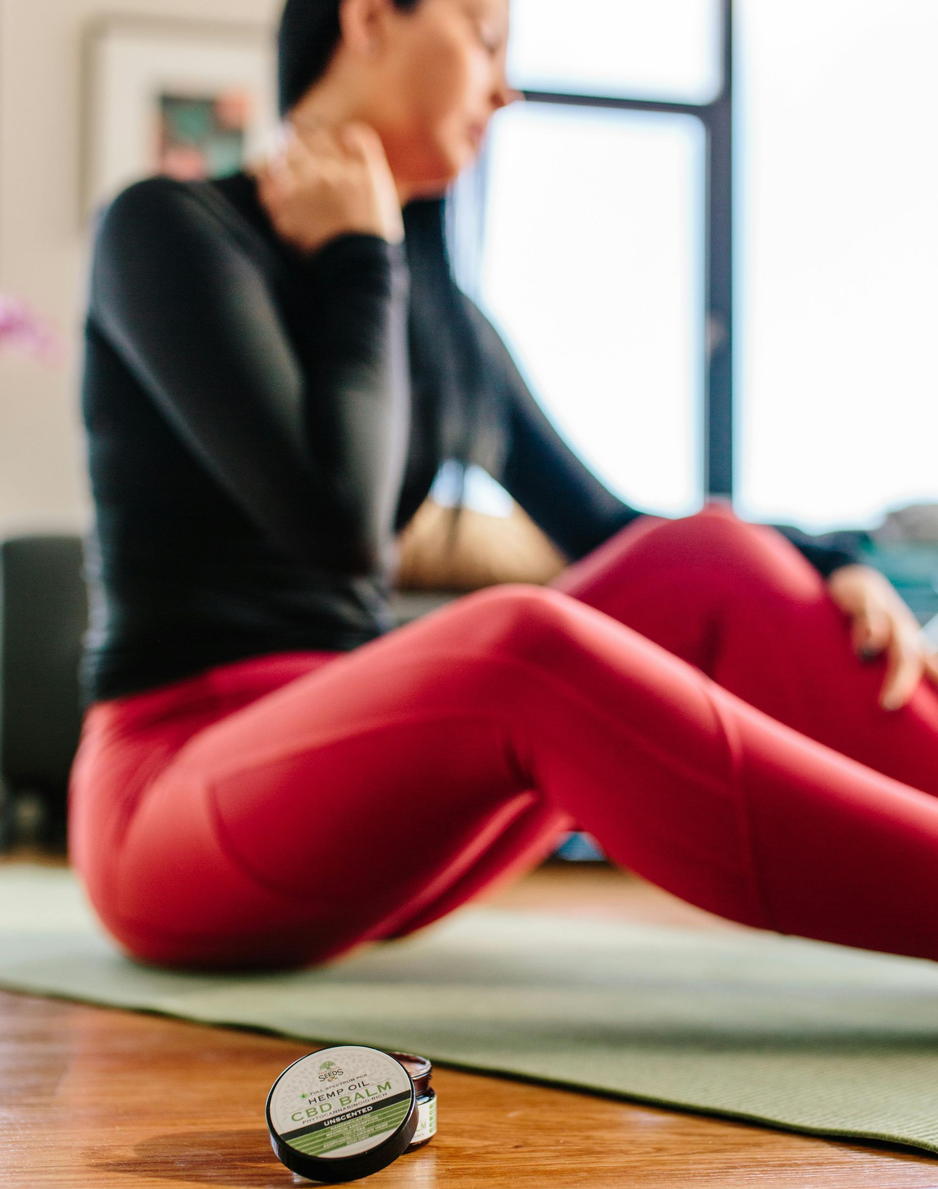 Woman in red leggings and black shirt sitting on mat, holding neck. Small product in foreground.