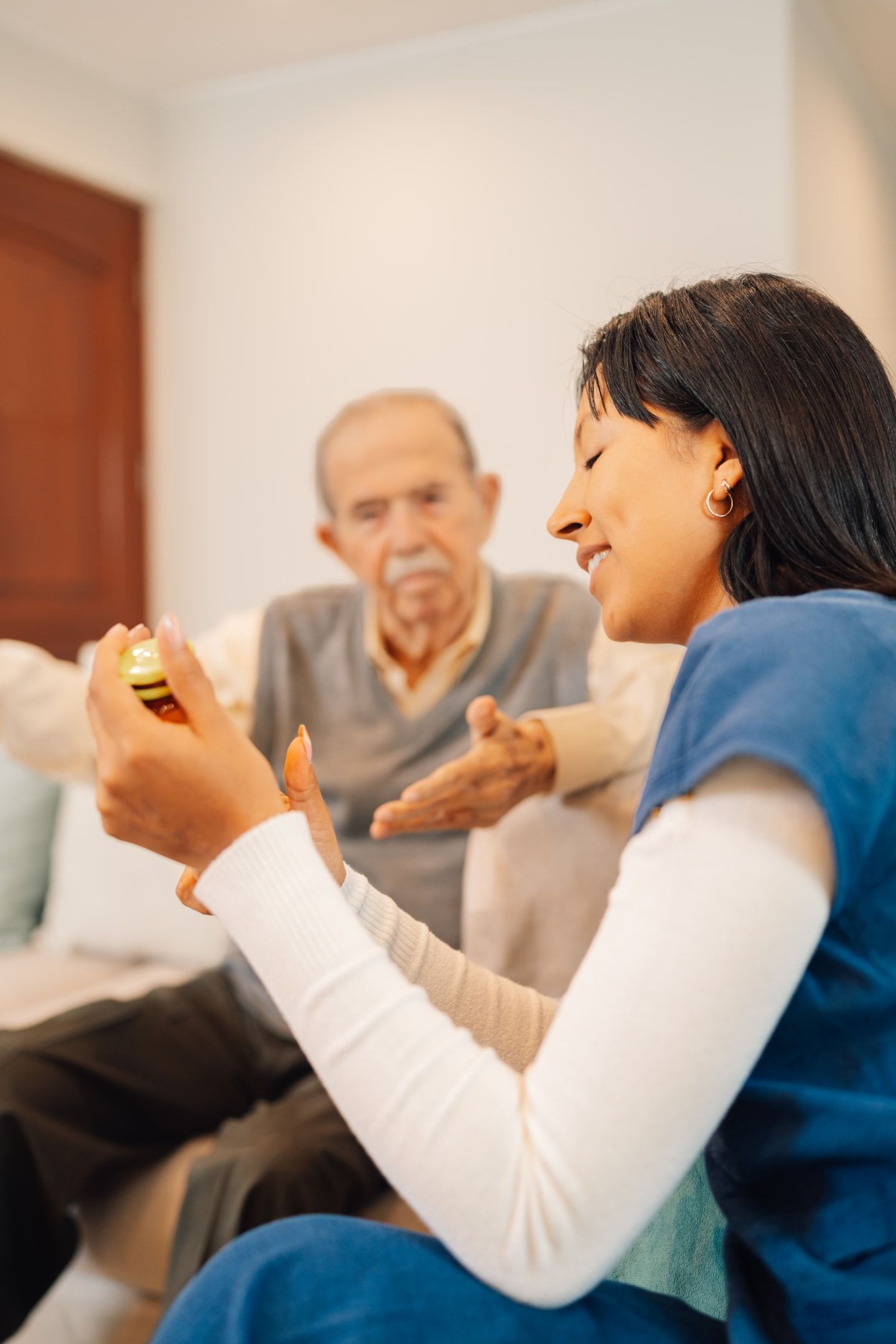 A healthcare worker shows medication to an elderly man, both seated indoors. The worker is smiling.