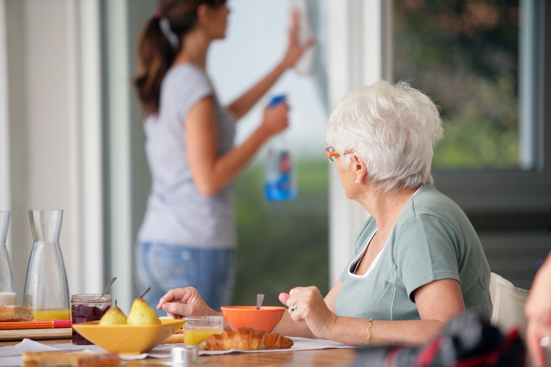 A woman is cleaning a window while an elderly woman sits at a table eating breakfast.