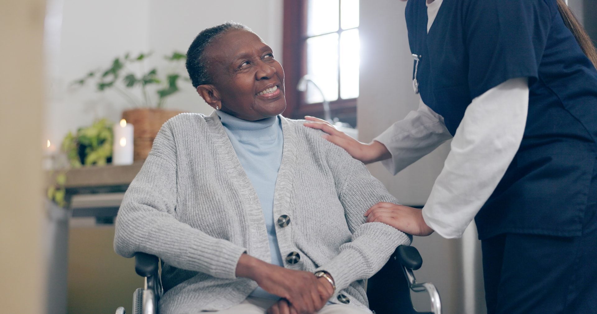 Older Black woman in wheelchair, smiling, with a healthcare worker's hand on her shoulder. Indoor setting.