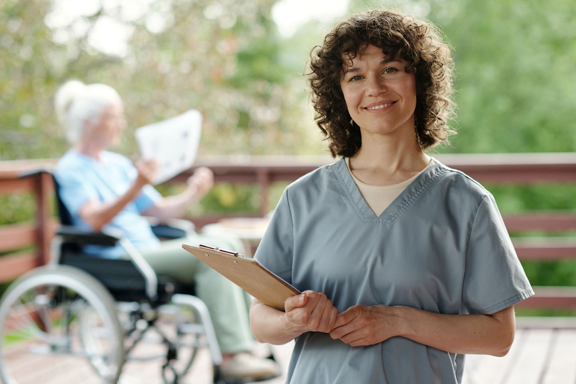 Caregiver in blue scrubs smiles, holding a clipboard, with a woman in a wheelchair reading in the background.