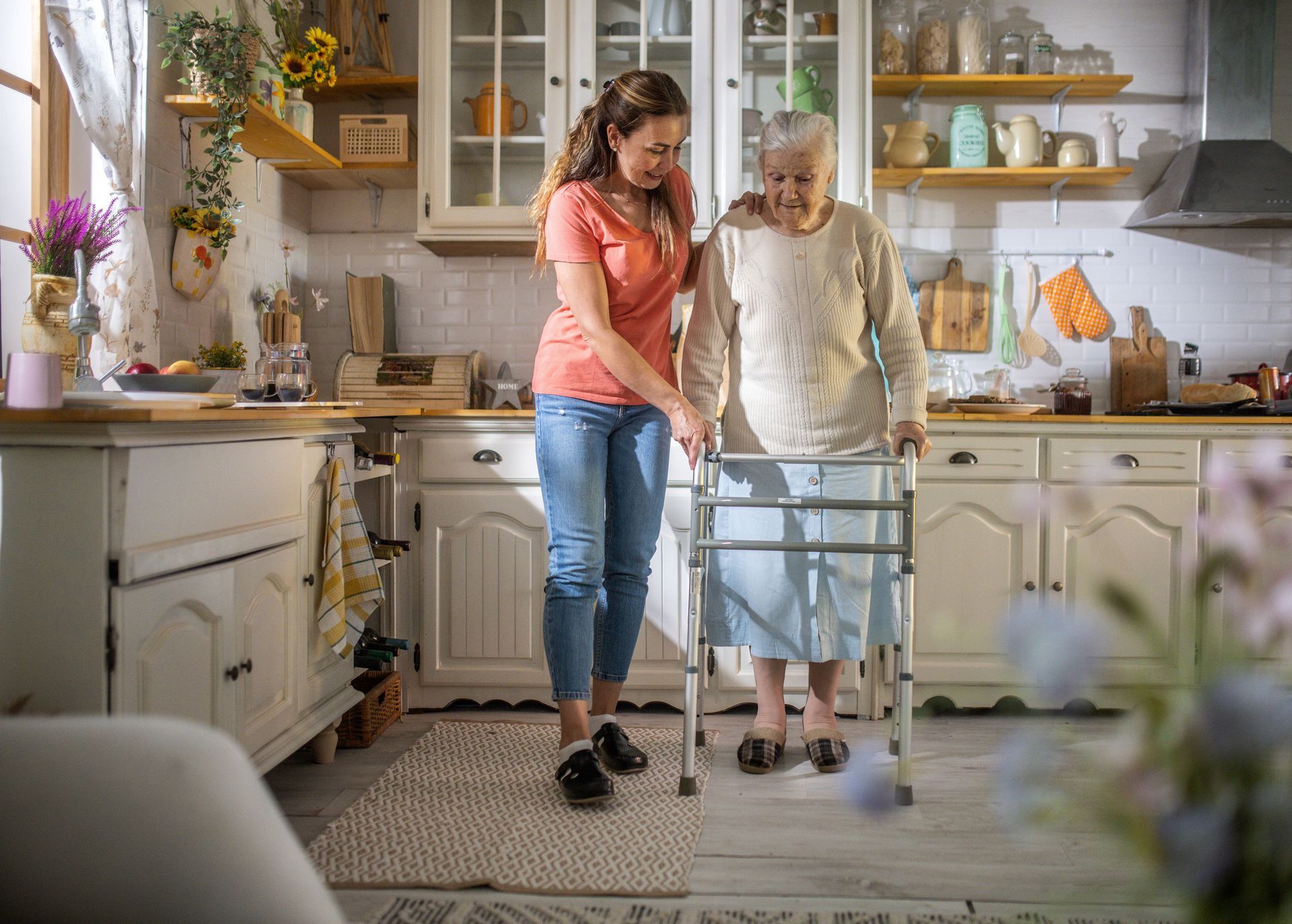 Woman helping elderly woman with walker in a bright kitchen.