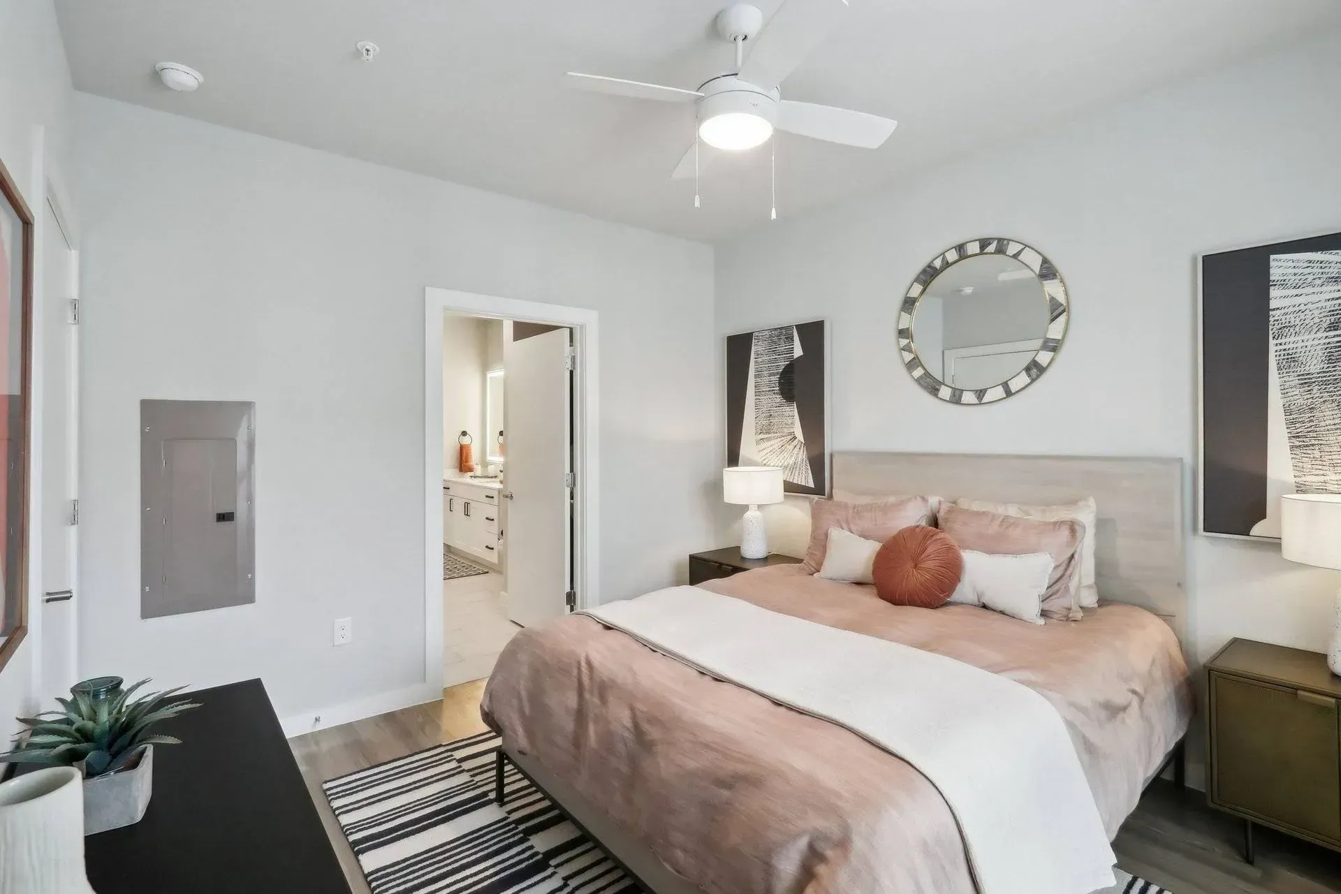 Bedroom with bed, rug, desk, artwork, and a doorway to a bathroom; light gray walls, warm tones.